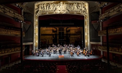 Sala de Espetáculos do Theatro Municipal com o palco iluminado no centro. No palco estão os membros e o regente da Orquestra Sinfônica Municipal. Nas laterais e estão três andares de poltronas e na parte inferior algumas pessoas assistindo.