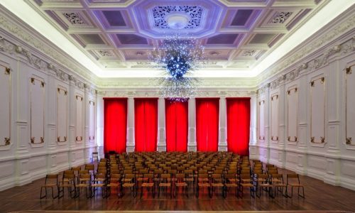 Uma sala branca, com paredes ornamentais, muitas cadeiras dispostas ao meio e um lustre grande azul na parte central da sala